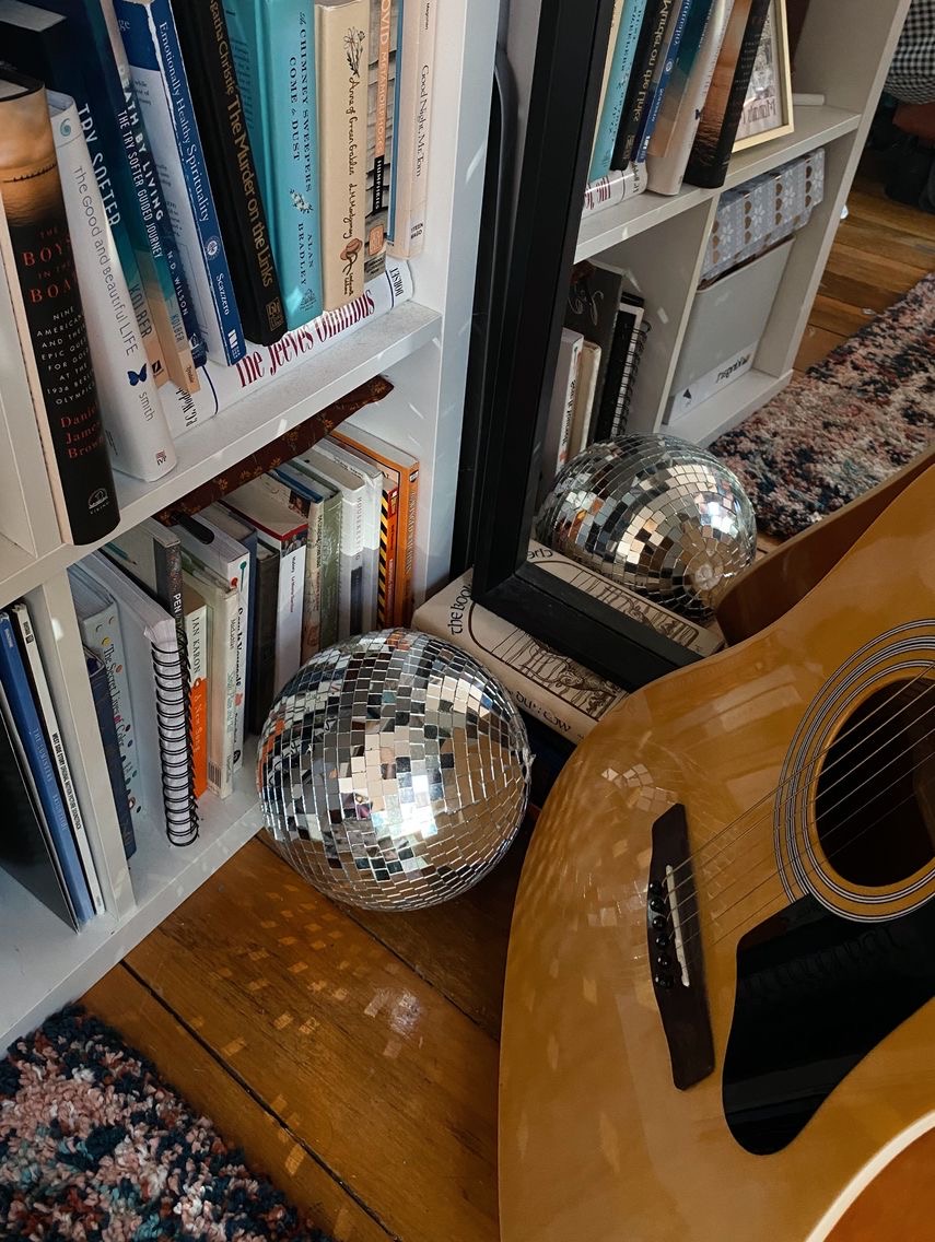 Estante de livros com um globo de discoteca no chão ao lado da estante, e um violão. Post "por que pessoas me associam a essas músicas?"