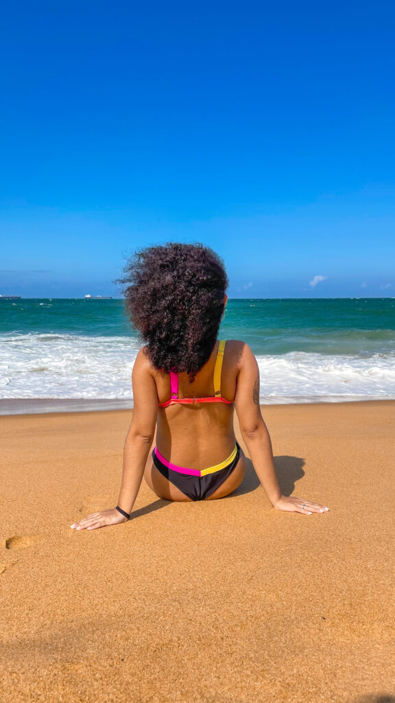 Foto minha na praia, sentada, de costas para a câmera e de frente pro mar